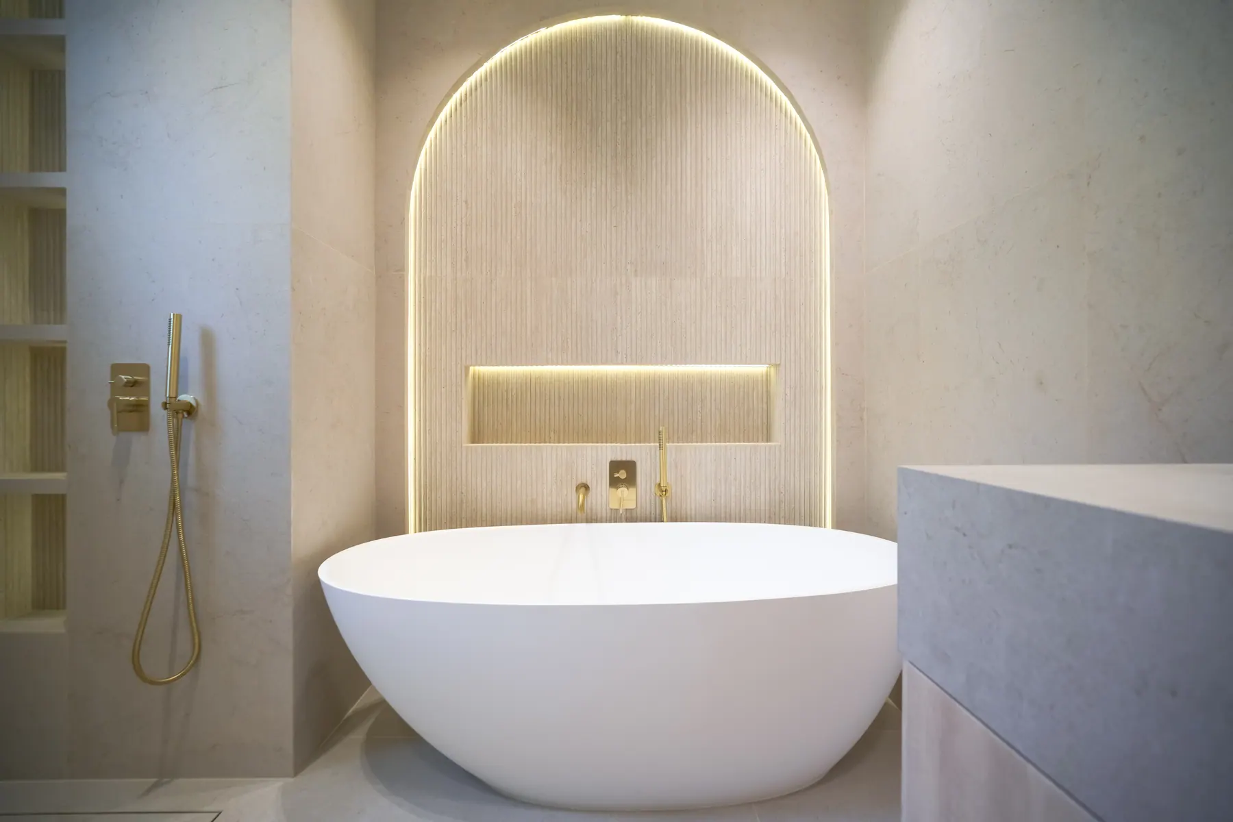 Travertine bathroom with a backlit fluted arch and a freestanding stone tub